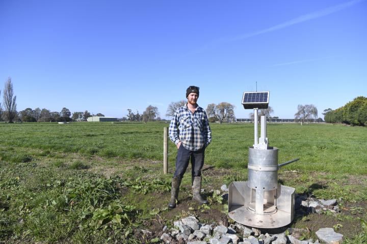 A man standing in a lush green farmland next to an irrigation water outlet, smiling at the camera. He is wearing gumboots, blue pants, and a blue and white checked shirt. 