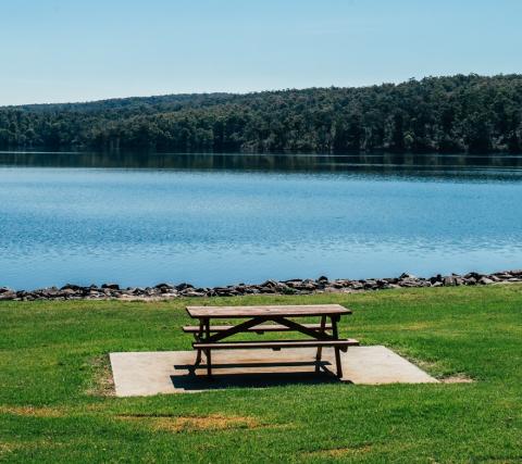 Picnic table on green grass, in front of a blue lake surrounded by trees